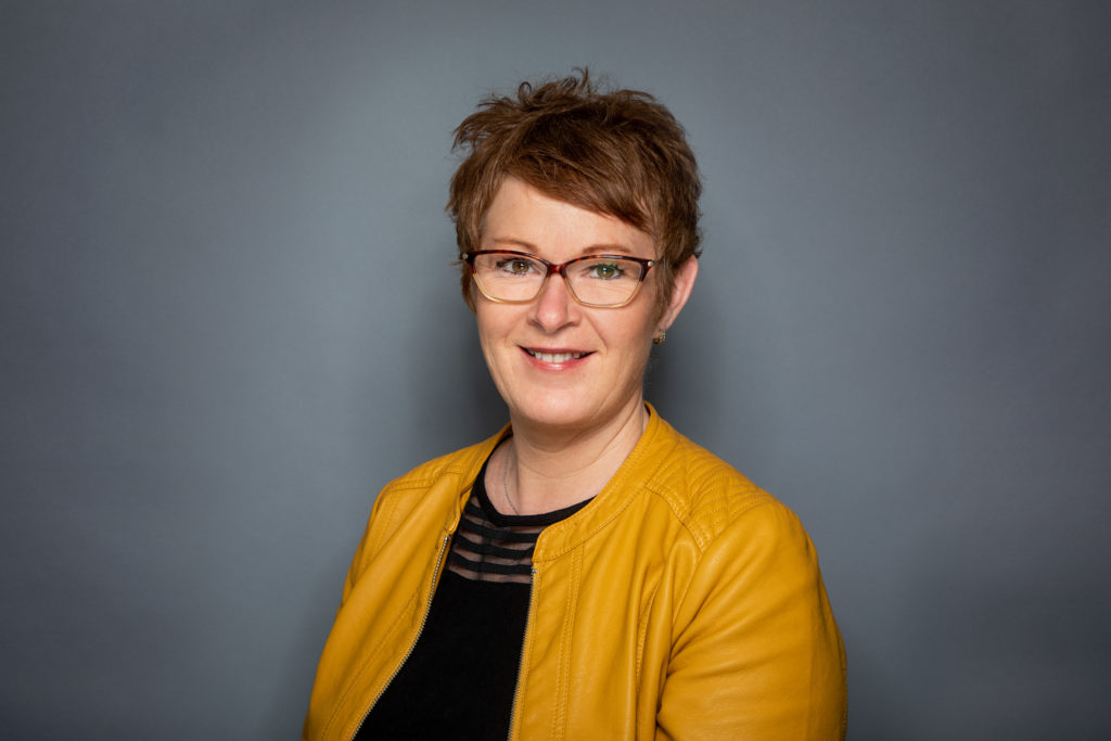 Portrettbilde av Solfrid Bøe, leder for arveutvalget i Norges Innsamlingsråd. Hun har mørkt, kort hår og briller. På seg har hun en gul jakke og sort topp.