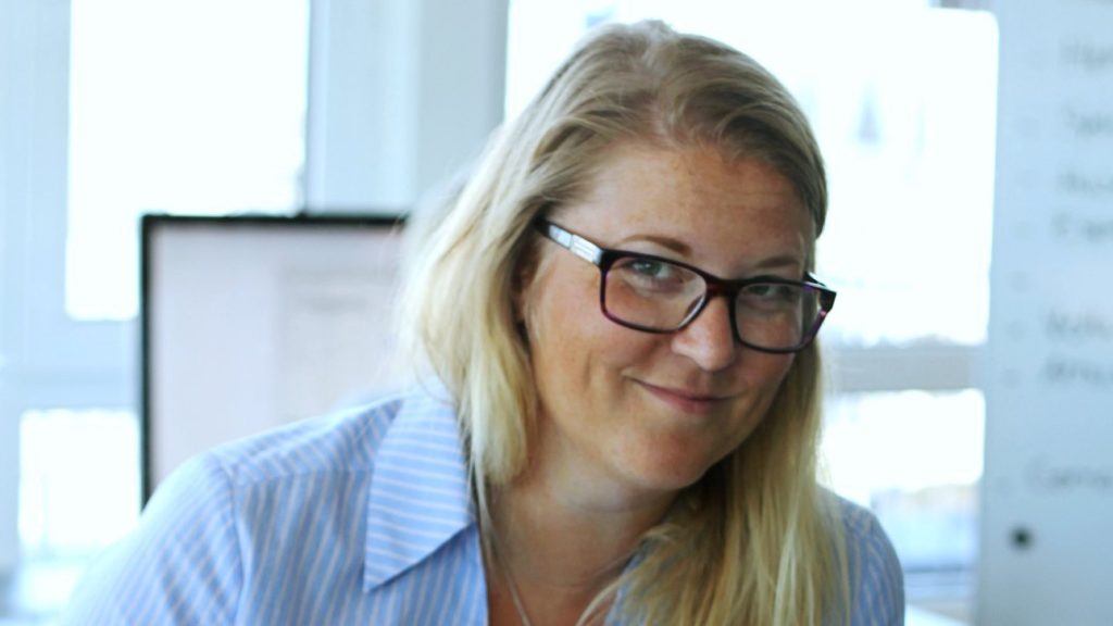Portrettbilde av Ane Tollerød Fosse. Hun har på seg mørke briller og en lyseblå skjorte. Hun sitter i et kontorlandskap.