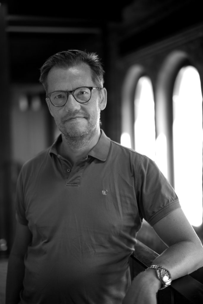 Portrettbilde av Klaus Damlien, styreleder i Fundraising Norge. Han har kort hår og bruker mørke briller. Klaus står foran et vindu. Bildet er i stort/hvitt.