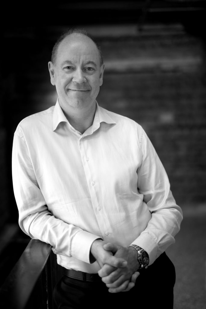 Portrettbilde av Knut Nordenhaug, styremedlem i Fundraising Norge. Han har på seg en hvit skjorte og mørk dressbukse. Knut står foran et vindu. Bildet er i stort/hvitt.