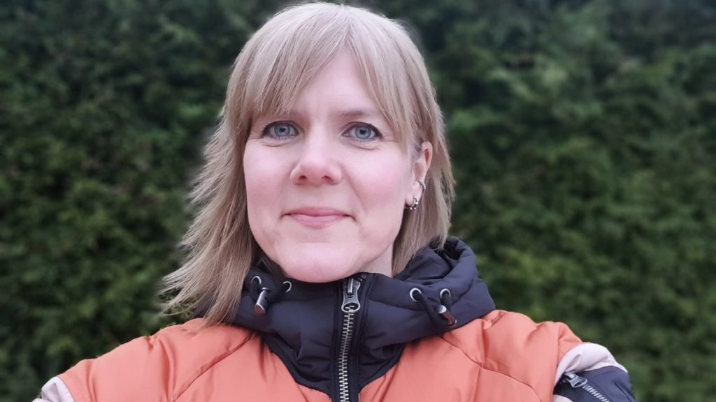 Potrettbilde av Trude Håskjold, spesialrådgiver i forretningsanalyse i Redd Barna. Hun har på seg en jakke i oransje og grått, og står ute i hagen.
