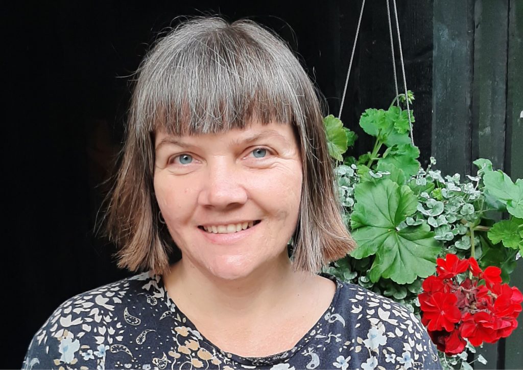Stina Neergård, fundraiser i Det Norske Misjonsselskap. Hun står mot en mørk husvegg, og har på seg blomstrete overdel. 